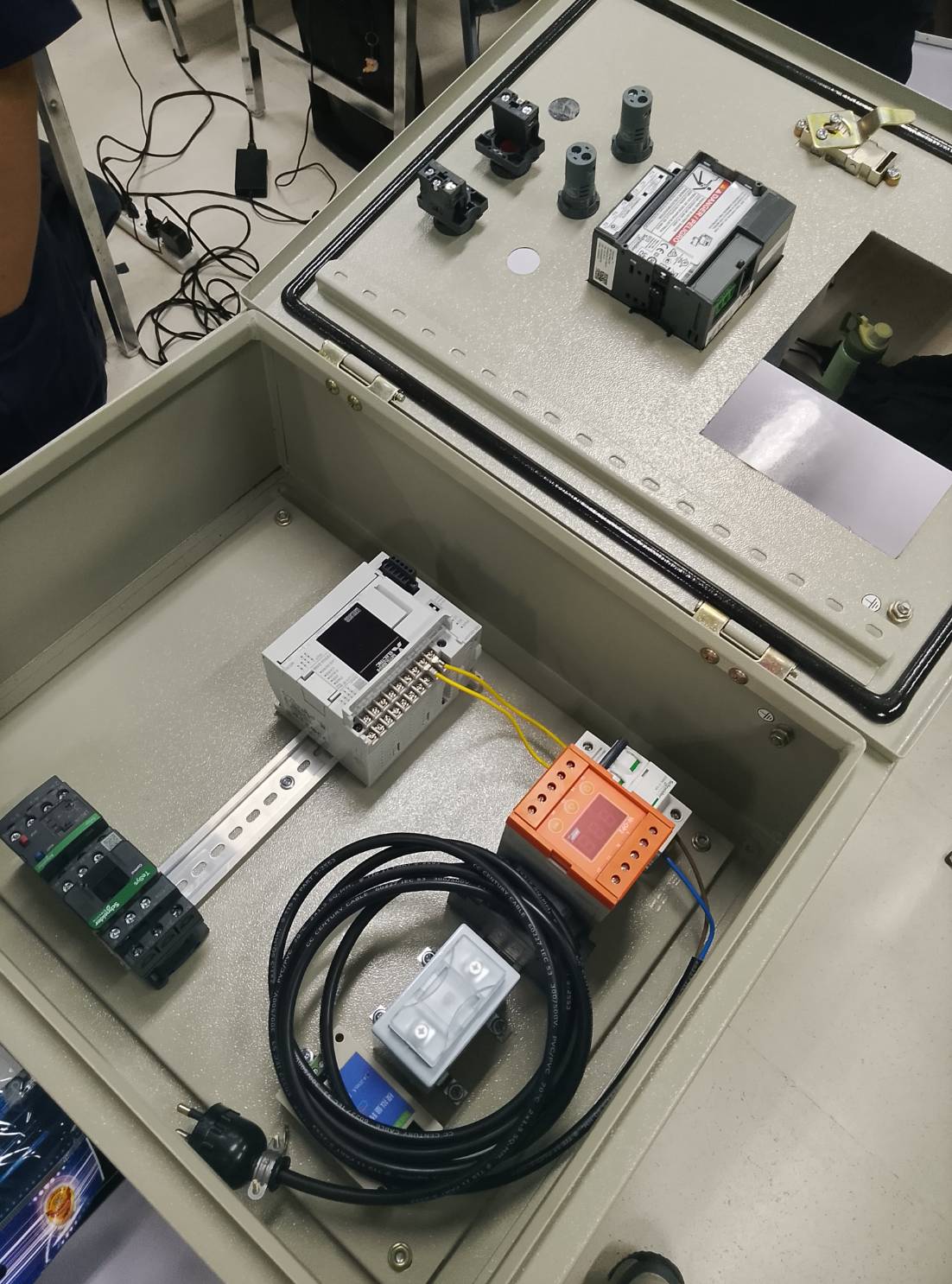 Open industrial control cabinet containing a PLC, circuit breaker and protection relay mounted on a DIN rail